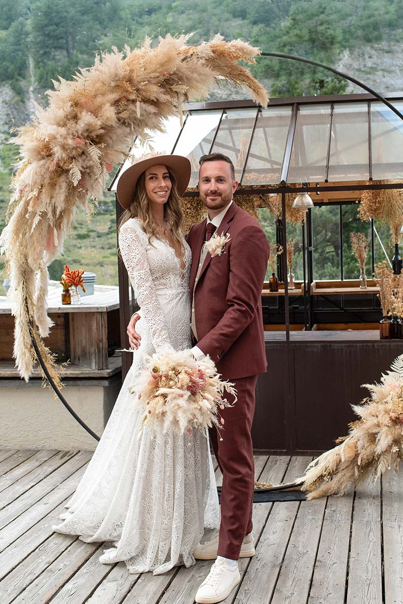 Boho-Brautpaar mit Hut unter einem Traubogen aus Pampasgras vor einem Gewächshaus